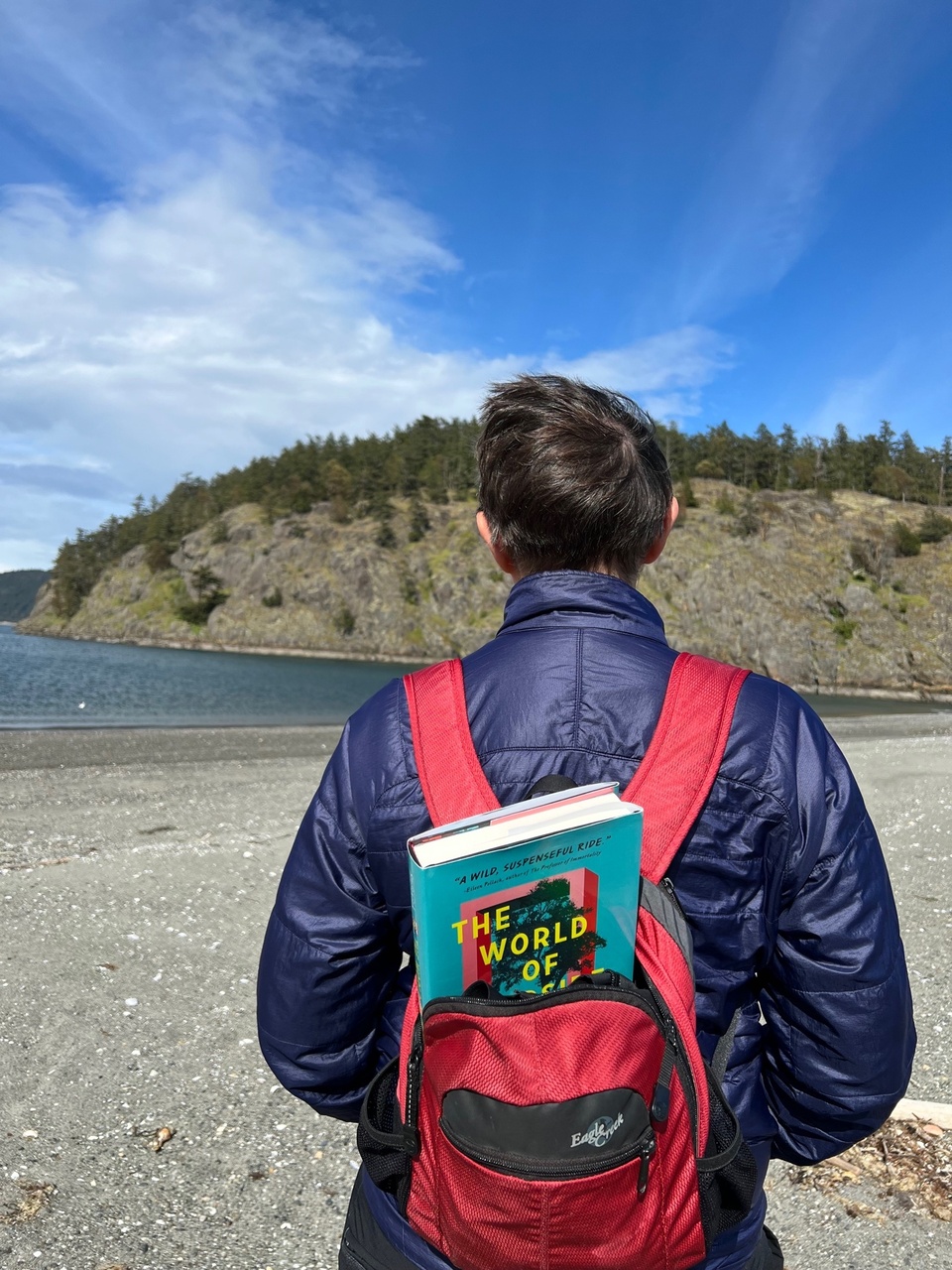 The World of Pondside book is peeking out of the backpack of a woman hiking near the ocean